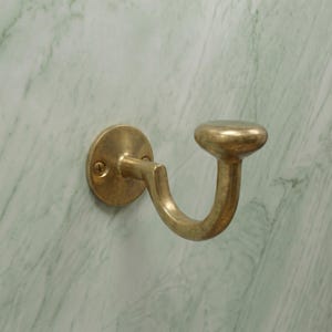 Pode incluir: Um gancho de latão montado na parede com uma placa de base circular e duas cabeças de parafuso visíveis. O gancho tem um braço curvo que se estende para fora e termina num botão arredondado em forma de cogumelo para pendurar objetos.