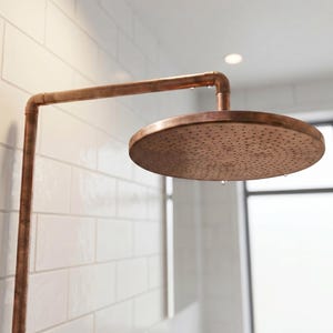Op de afbeelding: Een ronde koperen douchekop bevestigd aan een bijpassende koperen pijparm. De douchekop heeft een plat, cirkelvormig ontwerp met talloze kleine wateruitlaatopeningen over het oppervlak, met een metalen afwerking en subtiele textuur.