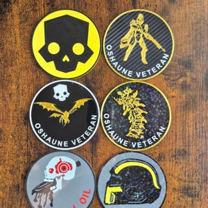 May include: A collection of six round stickers with various designs. The stickers feature images of skulls, a soldier, a spine, and a helmet. The text "OSHAUNE VETERAN" and "SPILL OIL" are also present on the stickers. The stickers are on a wooden surface.