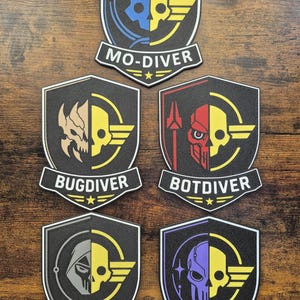 May include: Five black and white patches with a shield design. Each patch features a skull graphic, a wing, and a banner with text. The text includes "MO-DIVER", "BUGDIVER", "BOTDIVER", "STEALTHDIVER", and "SQUID-DIVER".