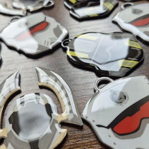May include: A collection of robot-themed charms and pendants in metallic grey, white, black, and red. Some have yellow accents and small loops for attaching. They are arranged on a wooden surface.