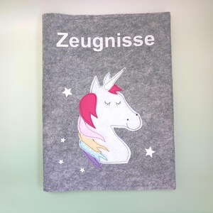 May include: A gray felt cover with the word "Zeugnisse" in white. A white unicorn with a pink mane and a rainbow tail is on the cover, along with white star accents. The cover is for documents.