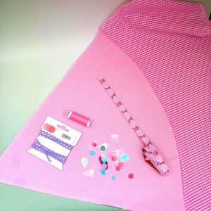 May include: A pink fabric surface with sewing supplies. Includes a spool of pink thread, purple lace trim, a patterned ribbon with a repeating design, and colourful confetti. A pink and white gingham fabric is in the background.