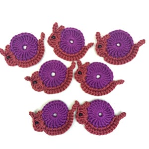 Op de afbeelding: Zeven handgemaakte gehaakte slakkenapplicaties. Elke slak heeft een paarse schelp en een roodbruin lichaam. De slakken zijn gerangschikt op een witte achtergrond, waarbij de details van het garen en het grillige ontwerp worden getoond.