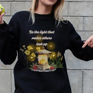 Może przedstawiać: Czarna bluza z napisem "Be the light that makes others look up." Projekt przedstawia akwarelową ilustrację latarni, kwiatów i liści. Osoba trzyma zielone jabłko.