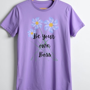 Op de afbeelding: Een lila T-shirt met een bloemenontwerp en de tekst "Be your own Boss" in zwart schrift. De bloemen zijn lichtblauw met gele centra en groene stelen.