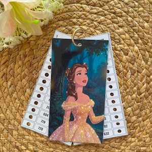 Puede incluir: Una tarjeta rectangular con la ilustración de una mujer de cabello castaño con un vestido amarillo, sujeta a una anilla metálica con tarjetas de inventario de papel blanco numeradas del 1 al 720, dispuestas en abanico sobre una superficie tejida.