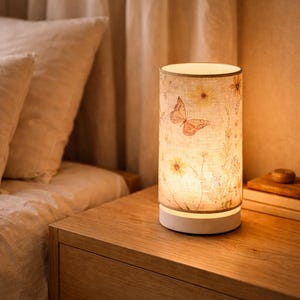 以下が含まれることがあります： 花と蝶のデザインが施された円筒形のベッドサイドランプ。 ランプは白いベースと、黄色い花と蝶で飾られたベージュのランプシェードを備えています。 木製のナイトスタンドの上に置かれています。