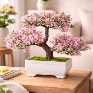 May include: A bonsai tree with pink flowers and green leaves in a white rectangular pot. The tree has a brown trunk and branches. The pot is on a wooden table. The table is made of wood.