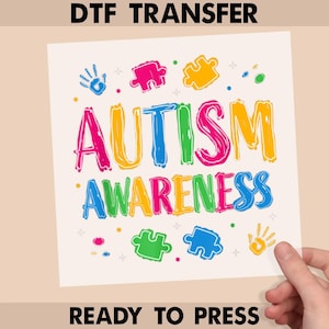 May include: A DTF transfer with the words "AUTISM AWARENESS" in colorful, outlined letters. The design includes puzzle pieces and handprints. The text "READY TO PRESS" is at the bottom.