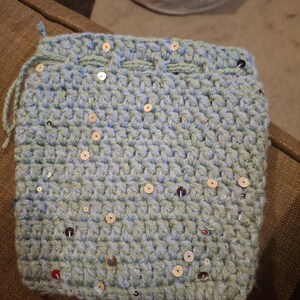 May include: A crocheted drawstring bag in a soft, light blue color, adorned with scattered silver sequins. The bag is handmade with a textured stitch, and the drawstring is a matching light blue yarn. The bag is a small size, perfect for carrying small items.
