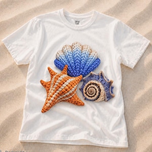 May include: White t-shirt with crochet seashell designs. The shirt features a blue and tan seashell, an orange and white starfish, and a blue and tan seashell. The text "SEA LIFE SERIES" is at the top.