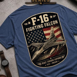 May include: Blue t-shirt featuring a graphic of an F-16 Fighting Falcon jet with an American flag design. The text on the shirt reads "F-16 Fighting Falcon" and "Peace Through Airpower 1974-Present". The shirt is part of the Aviation Series.