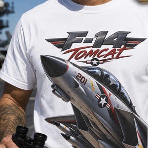 May include: A white short-sleeve t-shirt featuring a graphic of an F-14 Tomcat fighter jet with the text F-14 TOMCAT and AVIATION SERIES. The jet is silver with the number 201 and a star insignia. The shirt also includes the text Designed by Pixel2Life.