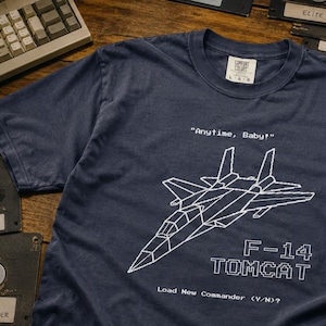 May include: A navy blue short-sleeve t-shirt featuring a white pixelated line drawing of an F-14 Tomcat fighter jet. The text reads "Anytime, Baby!", "F-14 TOMCAT", and "Load New Commander (Y/N)?". The shirt has a crew neck and a Comfort Colors label.