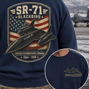 Op de afbeelding: Een marineblauw sweatshirt met een grote afbeelding op de achterkant van een SR-71 Blackbird vliegtuig boven een verweerd Amerikaans vlagschild. De tekst luidt SR-71 BLACKBIRD, STRATEGIC RECONNAISSANCE COMMAND, 1964-1998. Een kleine bijpassende afbeelding staat op de borst.