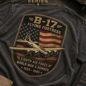 Może przedstawiać: Ciemnoszary t-shirt z grafiką samolotu B-17 Flying Fortress na tle postarzanej amerykańskiej flagi. Tekst zawiera napisy AVIATION SERIES, B-17 FLYING FORTRESS, EIGHTH AIR FORCE, WORLD WAR II BOMBER oraz 1938-1945.