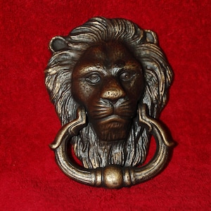 Könnte beinhalten: Türklopfer mit Löwenkopf in Bronzefarbe und Ringgriff. Das Gesicht des Löwen weist detaillierte Merkmale auf, darunter Mähne, Augen, Nase und Mund. Der Klopfer ist auf einem roten Hintergrund montiert.