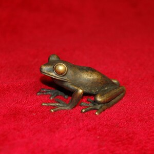 Puede incluir: Una figura de rana de bronce con grandes ojos dorados sobre una superficie roja. La rana está detallada con dedos visibles y una apariencia de piel texturizada. Es un artículo decorativo.