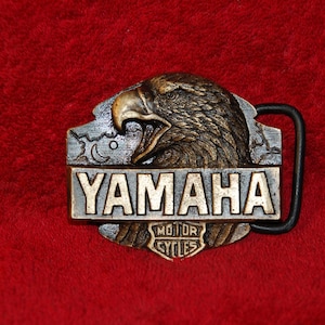 May include: A metal belt buckle featuring a detailed eagle head with open beak. The buckle has the Yamaha logo in a rectangular frame, with "Motor Cycles" below. The background is a deep red.