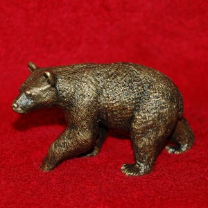 Puede incluir: Figura de oso de color bronce con textura de pelaje detallada, caminando sobre una superficie roja. El oso se representa en una pose realista, mostrando sus patas y la forma de su cuerpo. Artículo decorativo.