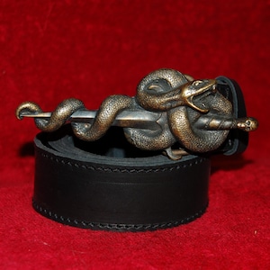 Pode incluir: Um cinto de couro preto com uma fivela de metal em tom de bronze em forma de serpente enrolada em torno de uma adaga. A serpente tem escamas detalhadas e a boca aberta, enquanto a adaga tem uma ponta afiada e um pequeno crânio no cabo.