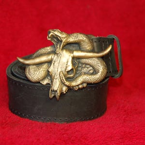 Könnte beinhalten: Ein schwarzer Ledergürtel mit einer großen, messingfarbenen Metallschnalle. Die Schnalle zeigt ein detailliertes, dreidimensionales Design einer gewundenen Schlange, die sich um einen Stierschädel mit markanten Hörnern legt, in einer texturierten Optik.