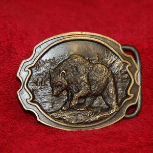 May include: Oval-shaped, bronze-colored belt buckle featuring a detailed relief of a bear in a natural landscape. The buckle has a decorative border and a sturdy clasp for securing a belt. The background is a deep red.
