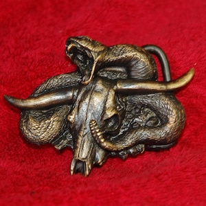 Puede incluir: Una hebilla de cinturón de color bronce que representa una calavera de toro con cuernos, entrelazada por una serpiente. La serpiente tiene la boca abierta y la hebilla está sobre un fondo rojo. La hebilla es probablemente de metal.