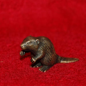May include: A bronze-colored figurine of a beaver, posed in a sitting position with its front paws raised. The beaver has detailed fur texture and a small tail. The figurine is set against a red background.