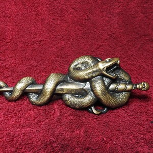 May include: A metallic bronze-toned belt buckle featuring a coiled snake wrapped around a horizontal dagger. The snake has an open mouth, and the dagger handle is topped with a small, detailed skull. The finish has a weathered, antique appearance.