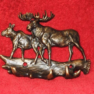 May include: A bronze-colored wall hook featuring a sculpted scene of two moose standing on a textured log base. The design includes three downward-curving hooks at the bottom and two circular mounting holes positioned on the log section.