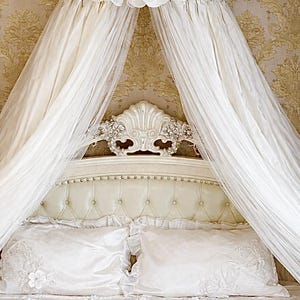 Könnte beinhalten: Ein luxuriöses weißes Bett mit Himmel. Das Kopfteil ist getuftet und mit einem dekorativen muschelförmigen Element verziert. Weißer, durchsichtiger Stoff drapiert vom Himmel. Die Bettwäsche umfasst bestickte Kissen und eine Bettdecke mit floralem Muster.
