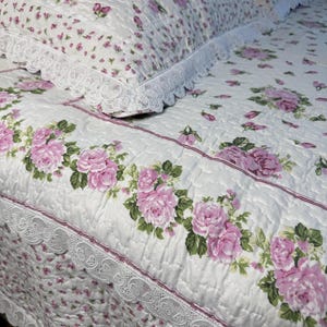 Pode incluir: Conjunto de roupa de cama acolchoada branca com um padrão floral de rosas em tons de rosa e verde. O conjunto inclui um edredom e fronhas, com acabamento em renda branca. O design é de estilo clássico e romântico.