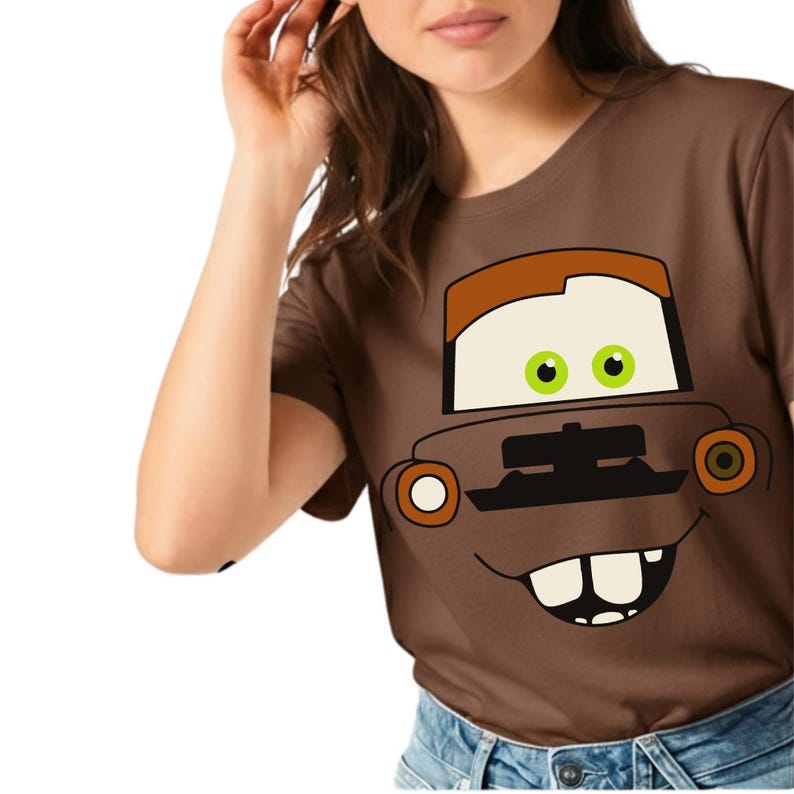 May include: Brown t-shirt featuring a cartoon car face with a brown top, off-white eyes, green pupils, and a smiling mouth. The car's design includes orange and white circular headlights. The shirt is made of a soft material.