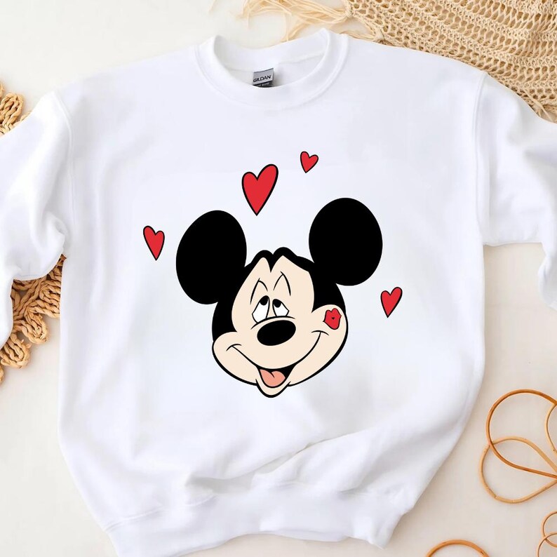 May include: White sweatshirt featuring a graphic of Mickey Mouse with red hearts and a kiss mark. The cartoon character has black ears, a smiling face, and is surrounded by floating hearts. A casual, comfortable top.