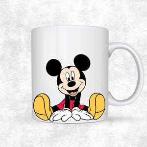 May include: White ceramic mug featuring a smiling Mickey Mouse illustration. The cartoon character is depicted in his classic outfit with black ears, red shorts, yellow shoes, and white gloves. The mug has a curved handle.