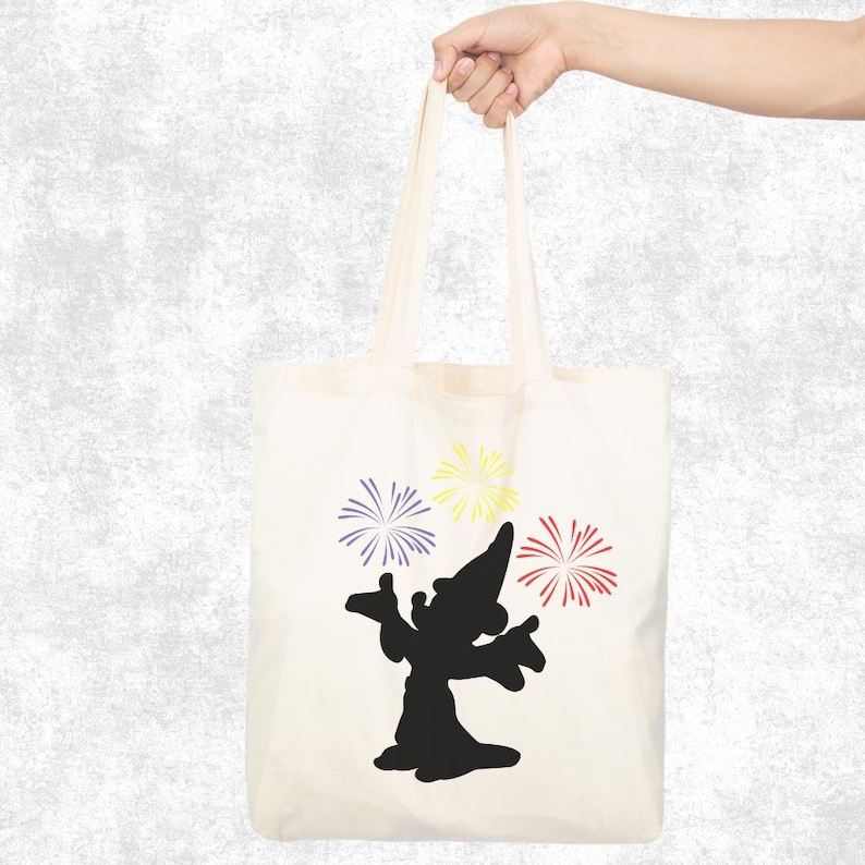 May include: Cream-colored tote bag featuring a black silhouette of a wizard with outstretched arms, accompanied by purple, yellow, and red fireworks. The bag is held by a hand against a textured gray background.