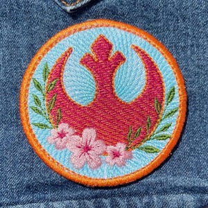 May include: A circular embroidered patch featuring a magenta Rebel Alliance symbol centered on a light blue background. The design is framed by an orange border and decorated with green leafy branches and three light pink cherry blossom flowers.