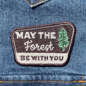 May include: A rectangular embroidered patch attached to blue denim fabric. The patch features a dark brown background with white text that reads MAY THE Forest BE WITH YOU and a green embroidered pine tree icon on the right side.