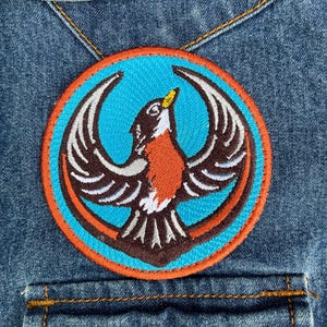May include: A circular embroidered patch featuring a brown and white bird with wings spread wide, set against a bright blue background. The design is framed by a thick orange border and is stitched onto a blue denim fabric with visible seams.