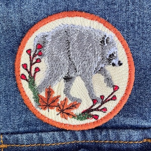 May include: A circular embroidered patch featuring a grey raccoon walking across a cream background. The design includes orange maple leaves and thin branches with small red berries, all enclosed by a vibrant orange stitched border on denim.