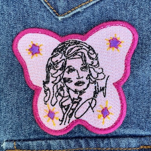 May include: A butterfly-shaped embroidered patch on blue denim fabric. The patch features a light pink background with a dark pink border and a black line-art portrait of a person with wavy hair. Four small purple stars with yellow rays surround the portrait.