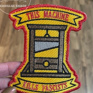 May include: Embroidered patch with a yellow background and a red border. The patch features a guillotine design with the text "THIS MACHINE" above and "KILLS FASCISTS" below in red lettering.