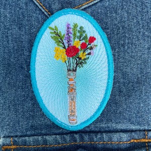 May include: An oval embroidered patch featuring a bouquet of colorful flowers in a tall, slender vase against a light blue background. The flowers include red, yellow, and purple blossoms with green stems. The patch has a bright cyan border.