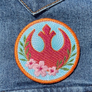May include: A circular embroidered patch featuring a dark pink Rebel Alliance symbol centered on a light blue background. The design is framed by a bright orange border and decorated with green leafy branches and three light pink cherry blossom flowers.