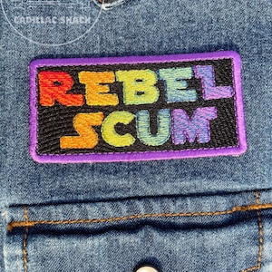 May include: A rectangular embroidered patch with the words "REBEL SCUM" in rainbow colors on a black background, framed by a purple border. The patch is affixed to a denim jacket.