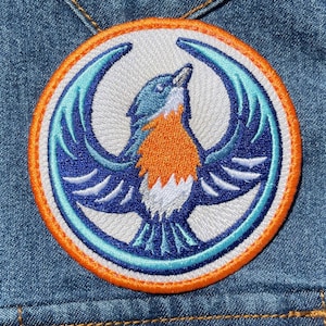 May include: Embroidered patch featuring a stylized bird with outstretched wings. The bird has a blue head and wings, an orange breast, and a white belly. The patch is circular, with a white background, blue accents, and an orange border. It is sewn onto a denim jacket.