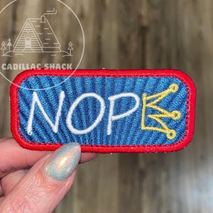 May include: Embroidered patch with the word "NOPE" in white, a gold crown, and a blue background with a striped pattern. The patch has a red border. The text "CADILLAC SHACK" is in a circle in the upper left corner.