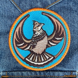 May include: A circular embroidered patch featuring a stylized bird with outstretched wings in shades of brown, tan, and white. The bird is centered on a bright blue background, enclosed by a vibrant orange border, stitched onto blue denim fabric.